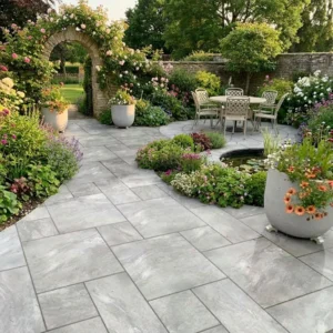 A stone patio with Quartz Grey Outdoor Porcelain Paving Tiles (Mixed Sizes) features a round table and chairs by a pond, surrounded by flowering plants, large pots, and an archway of climbing blooms leading to a grassy garden.