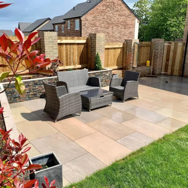 A modern backyard patio features cushioned wicker furniture on a tiled surface enhanced by Raj Green Outdoor Porcelain Paving Tiles (600mm x 900mm x 20mm). The area is enclosed by a wooden fence, accompanied by a small garden and brick accent wall, with lush green grass bordering the patio.
