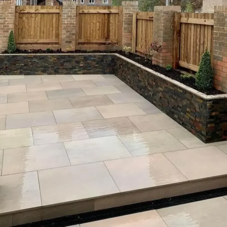 A modern patio features Raj Green Outdoor Porcelain Paving Tiles and a raised garden bed with greenery and small trees bordered by a stone wall. Enclosed by wooden fences and brick columns, the space is neat and clean.