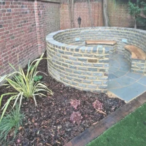 The garden features a curved stone seating area with wooden benches, encircled by Kandla Grey Sandstone Walling. Adjacent to this space is a landscaped area adorned with mulch and plants. Beyond the setting, a brick fence and trees enhance the background.