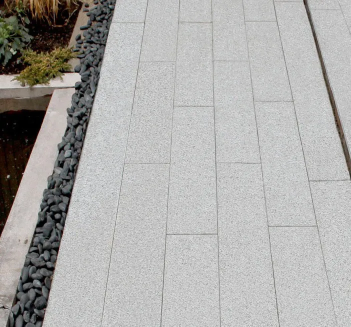 A modern patio features Silver Grey Granite G603 Planks (200mm x 900mm), bordered by smooth black river rocks and a small landscaped garden bed with greenery.