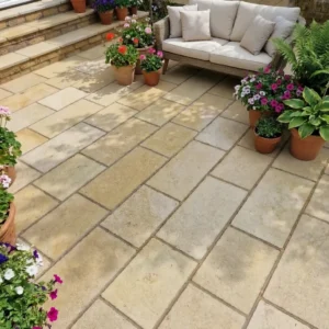 A stone patio with Yellow Limestone Sawn 22mm Calibrated 600mm x 900mm tiles features a wicker sofa with white cushions, surrounded by potted flowering plants and ferns. Steps lead up to another level in the background.