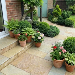 Terracotta pots with vibrant plants and flowers are displayed on a patio and steps made of Yellow Limestone Honed and Brushed - 22mm Calibrated- Sawn - Mixed Sizes, leading to a garden with manicured shrubs, trees, and a lush lawn.