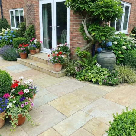 A patio of Yellow Limestone Honed and Brushed stones leads to glass doors, with steps lined by colorful plants, a ceramic urn among greenery, and blooming hydrangeas by the brick house.